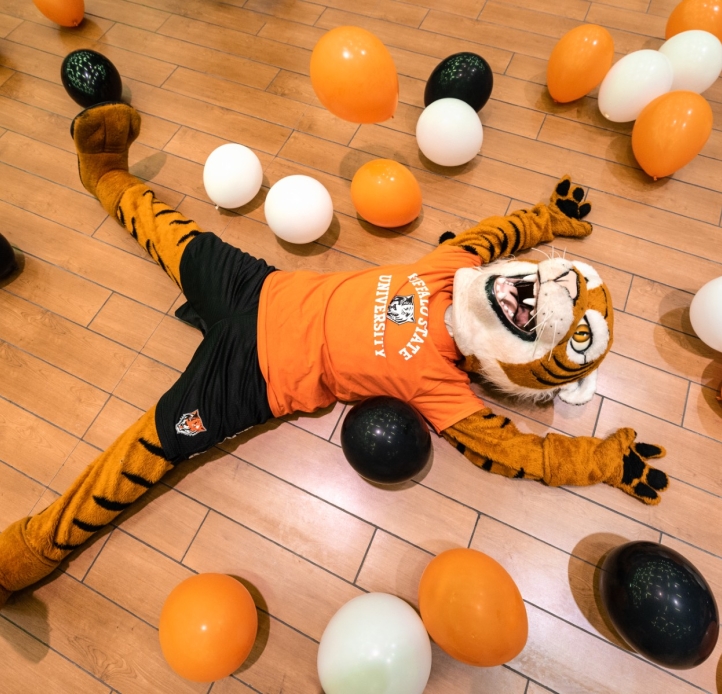 Benji laying on the ground surrounded by different colored balloons, orange, white and black